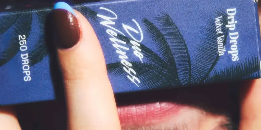 Close-up of a blue boxed product with a tropical palm leaf pattern, held by fingers with blue-painted nails; white cursive text is visible on the packaging.